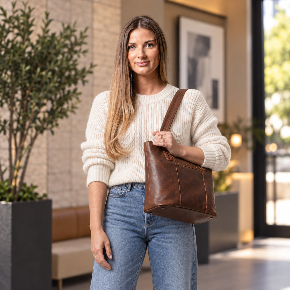 A woman wearing a cream ribbed knit sweater and blue jeans carries a brown leather tote bag with a single shoulder strap in a modern indoor setting. Concealed Carry Peyton Leather Tote for Women Lady Conceal - YKK Locking & Universal Holster