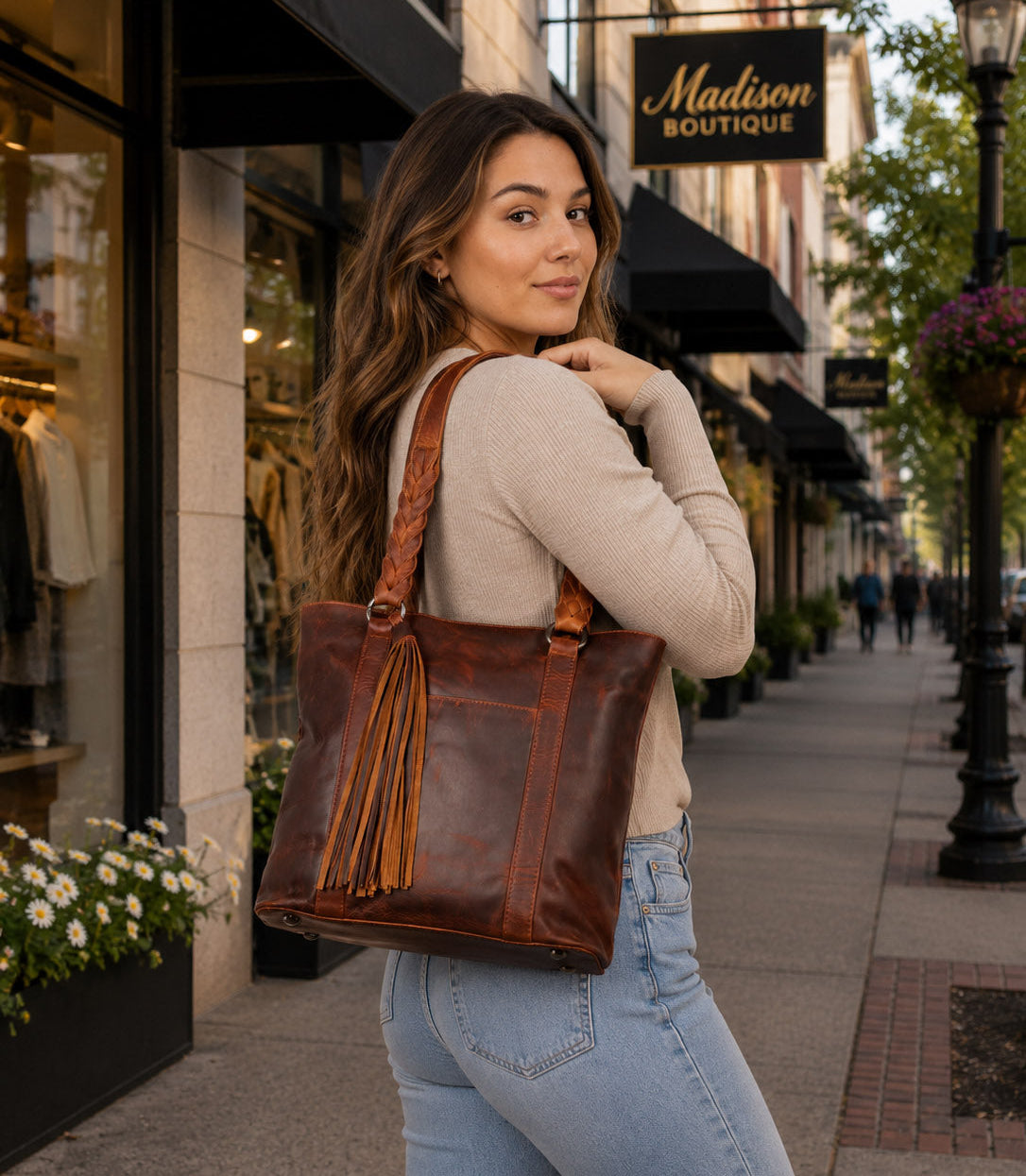 A woman wearing a beige long-sleeve top and light blue jeans carries a large brown leather tote bag with tassel fringe on a city sidewalk carrying The Bella Concealed Carry Tote  in Cognac