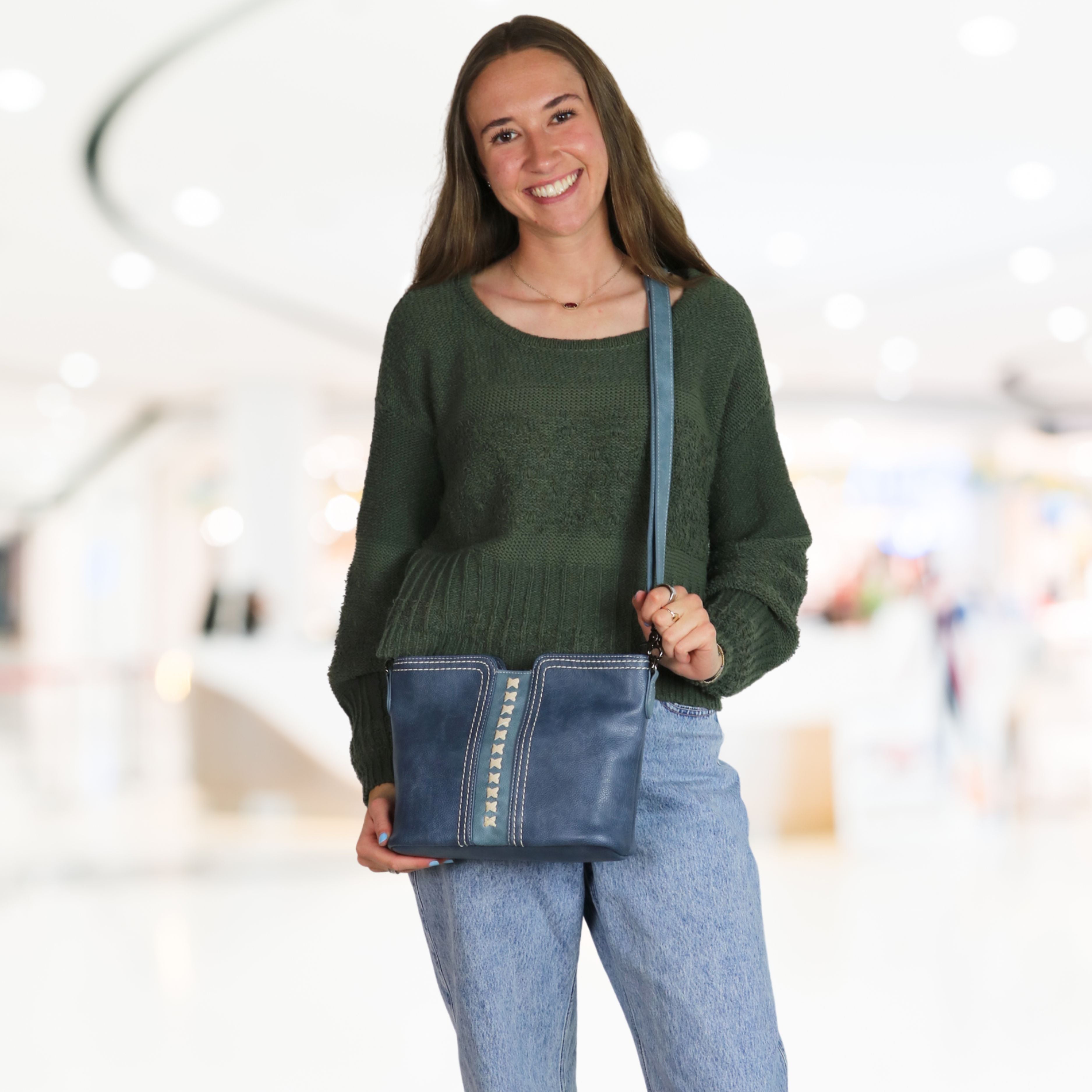 A woman wearing a dark green knit sweater and light blue jeans carries a blue leather crossbody bag with gold-tone studs and a blue strap in a brightly lit, blurred indoor shopping mall setting.