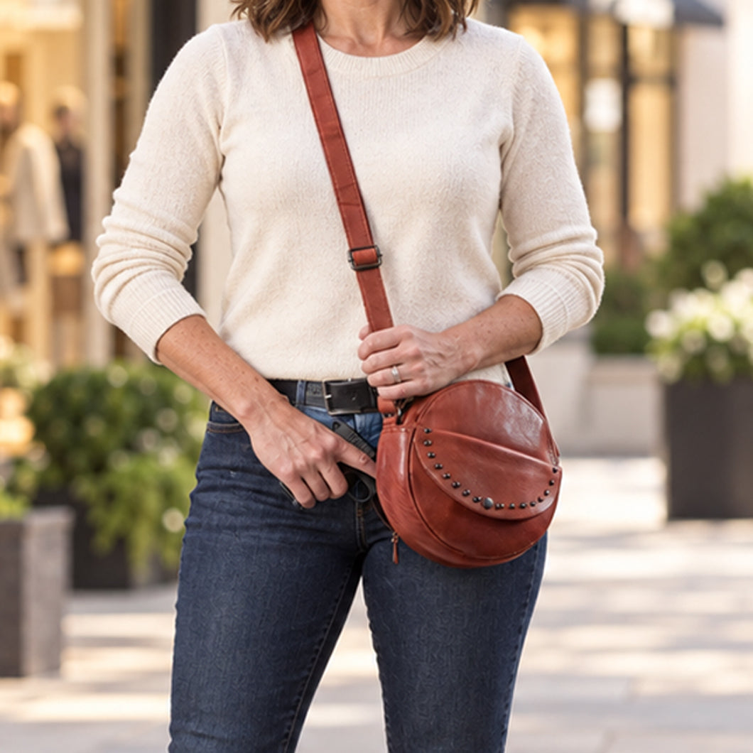 A woman wearing a cream knit sweater and blue jeans carries a brown leather crescent-shaped crossbody bag with a matching strap and decorative studs, photographed in an outdoor urban setting. Concealed Carry Mia Crossbody Purse -  Locking Zippers and Universal Holster Tactical Bag for Women  -  YKK Locking Gun Purse -  Concealment Pocket -  Pistol Women's Purse Crossbody