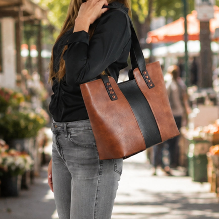 A woman wearing a black long-sleeve top and gray jeans carries a large brown faux-leather tote bag with a wide black center stripe and black shoulder straps in an outdoor street market setting.